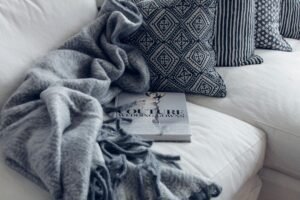 A stylish living room featuring a white sofa with patterned pillows, a blanket, and a fashion book.